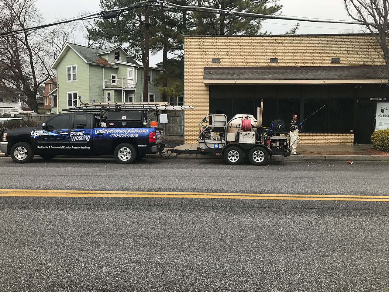 Professional pressure washing service truck and trailer parked outside a commercial building while technicians clean the exterior with specialized equipment.
